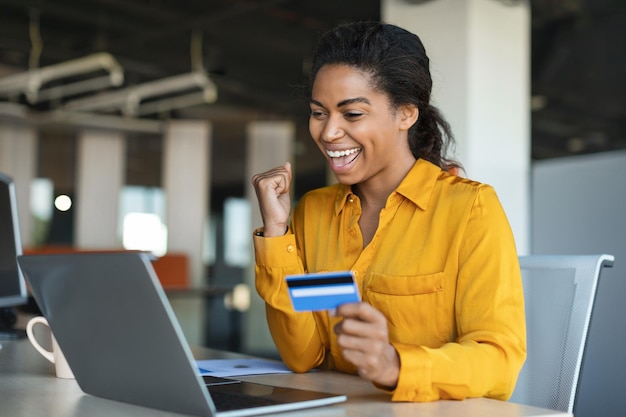Donna di affari afroamericana felicissima che fa gesto di successo tenendo la carta di credito seduta al tavolo con il computer portatile