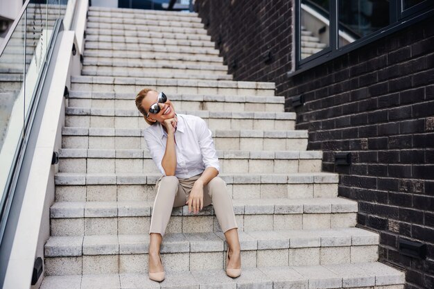 Donna di affari alla moda che si siede sulle scale davanti al centro d'affari e in posa