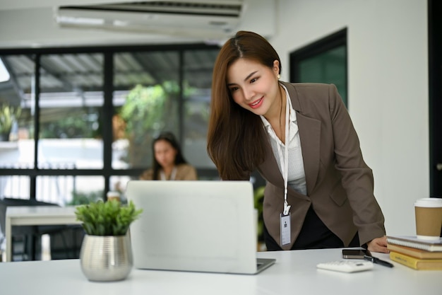 Donna di affari asiatica attraente che si appoggia sul tavolo e usando il suo computer portatile alla sua scrivania