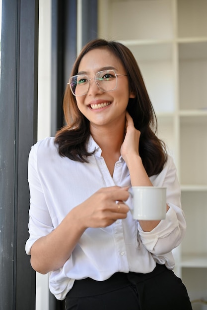 Donna di affari asiatica attraente che tiene una tazza di caffè che si appoggia sulla finestra