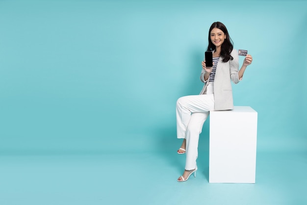 Donna di affari asiatica che si siede sulla scatola bianca e che mostra la carta di credito ed il telefono cellulare