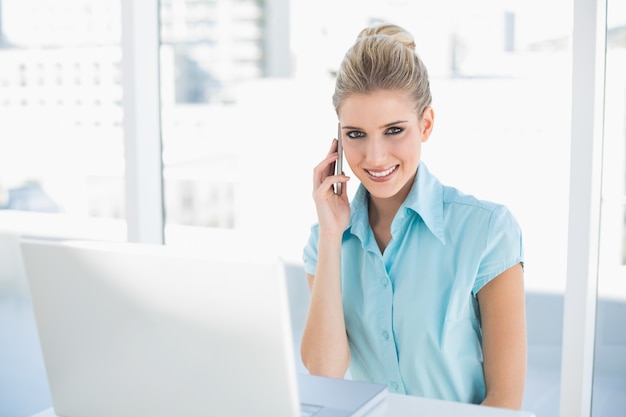 Donna di affari astuta pacifica sul telefono mentre per mezzo del computer portatile