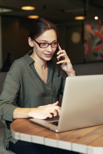 Donna di affari attraente in giacca nera e vetri alla moda che si siedono in caffè, parlando al telefono e guardando portatile.