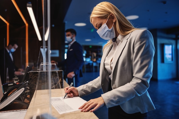 Donna di affari bionda con la maschera per il viso in piedi alla reception in hotel e riempire il modulo durante la pandemia del virus corona. Viaggio di lavoro, viaggio durante la corona, misure precauzionali COVID19