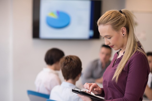 Donna di affari bionda in abiti casual che lavora su computer tablet all'interno di un moderno ufficio affari di avvio. Gruppo di giovani sulla riunione di squadra sfocata sullo sfondo