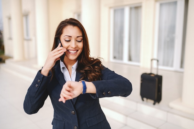 Donna di affari caucasica sorridente dei giovani che esamina orologio e che per mezzo dello Smart Phone mentre stando all'aperto. Nel bagaglio di fondo. Concetto di viaggio d'affari.