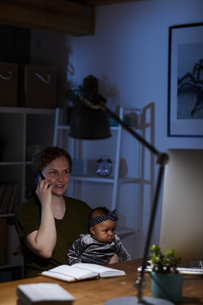 Donna di affari che lavora a casa fino a notte che parla al telefono cellulare mentre era seduto al tavolo con la sua bambina