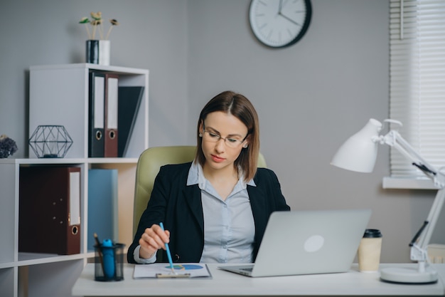 Donna di affari che lavora ad un computer portatile nel suo rapporto finanziario moderno della carta della tenuta dell'ufficio.