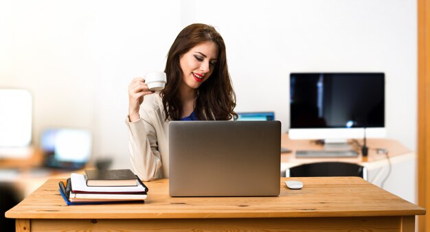 Donna di affari che lavora con il suo computer portatile e che tiene una tazza di caffè nell&#39;ufficio