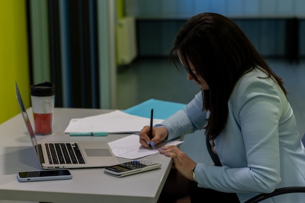Donna di affari che scrive con la matita sul blocco note in ufficio con il computer portatile e la calcolatrice sul tavolo