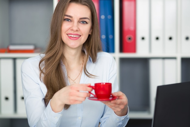 Donna di affari che tiene una tazza di caffè rossa in sue mani. Pausa al lavoro. Guarda la telecamera e sorride.