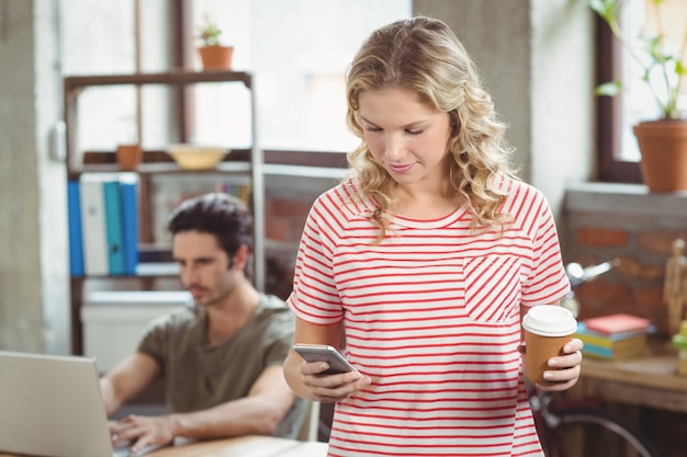 Donna di affari che utilizza smartphone mentre tenendo caffè nell&#39;ufficio luminoso creativo