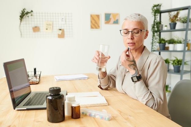 Donna di affari dai capelli corti in camicia che prende la pillola e la beve con l'influenza dell'acqua degli integratori o