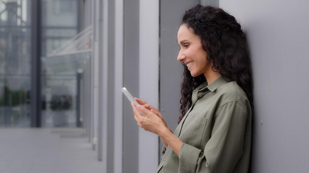 Donna di affari della donna ispanica caucasica di vista laterale che tiene telefono cellulare che sta vicino alla costruzione della parete