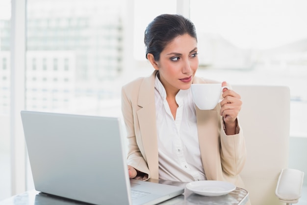 Donna di affari di pensiero che tiene tazza mentre lavorando al computer portatile