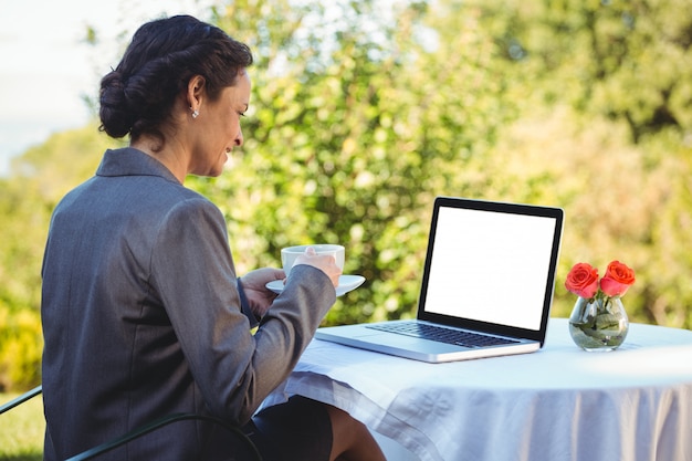 Donna di affari graziosa che mangia caffè e che per mezzo del computer portatile