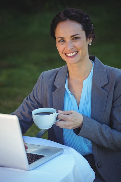 Donna di affari graziosa che mangia caffè e che per mezzo del computer portatile