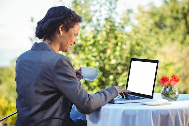 Donna di affari graziosa che per mezzo del computer portatile e mangiando caffè