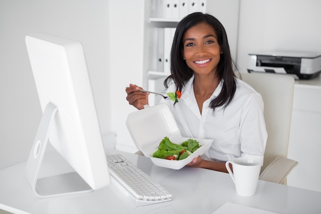 Donna di affari graziosa felice che mangia un&#39;insalata al suo scrittorio