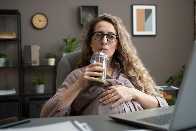 Donna di affari incinta che beve frullato mentre si siede davanti al computer portatile