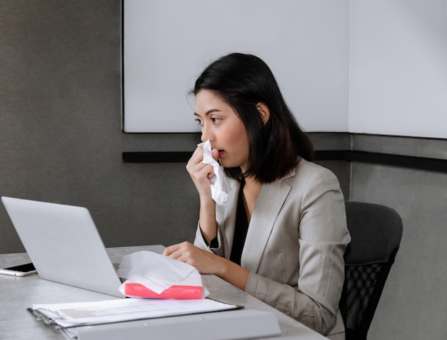 Donna di affari malata che si siede alla scrivania con il computer portatile, starnutendo e tossendo