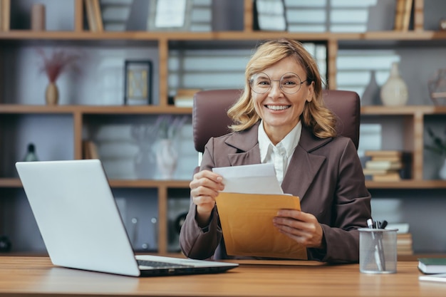 Donna di affari matura sorridente con gli occhiali che lavora al computer portatile e che tratta documenti in a