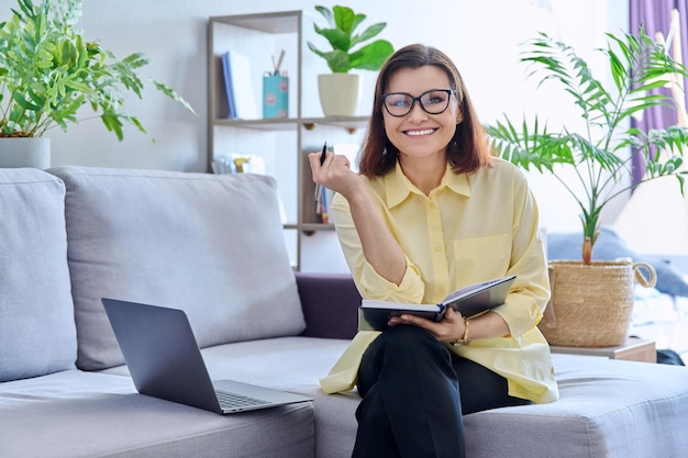Donna di affari maturi che lavora sul divano utilizzando il computer portatile che guarda l'obbiettivo
