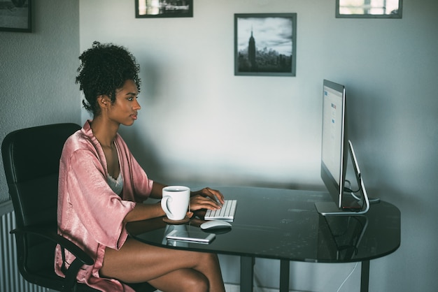 Donna di colore a casa che lavora con il computer e il caffè di mattina