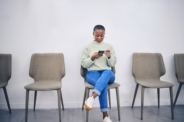 Donna di colore al telefono e seduta su una sedia in una sala d'attesa con connessione internet per i social media Persona di sesso femminile africana in fila per il colloquio di reclutamento con uno smartphone per l'app di comunicazione