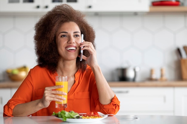 Donna di colore allegra che parla al cellulare e fa colazione in cucina