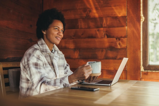 Donna di colore che beve il tè del mattino e usa il computer portatile mentre trascorre le vacanze in un'accogliente casa di legno.