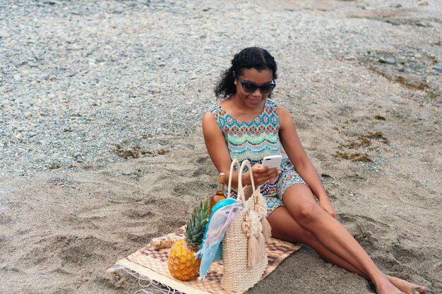 Donna di colore che controlla il suo cellulare seduta sulla spiaggia