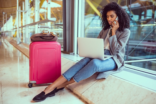 Donna di colore che per mezzo del telefono cellulare e del computer portatile all'aeroporto che si siede alla finestra