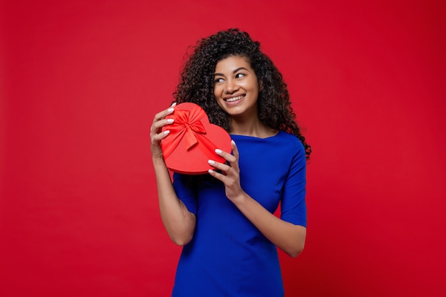 Donna di colore che sorride con il contenitore di regalo a forma di cuore isolato sulla parete rossa