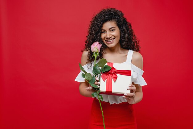 Donna di colore civettuola allegra con la rosa di rosa e presente romantico isolato sopra rosso