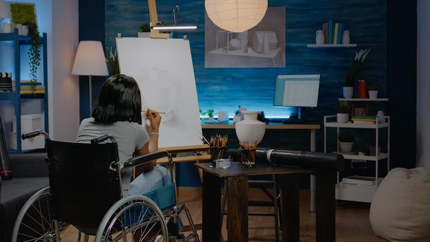 Donna di colore con disabilità disegno vaso su tela utilizzando strumenti artistici e matita. Giovane artista afroamericano in sedia a rotelle che progetta un capolavoro moderno per un progetto innovativo di belle arti