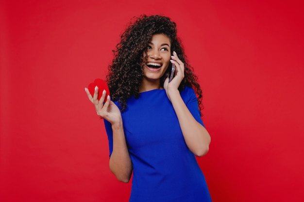 Donna di colore con la carta del biglietto di S. Valentino a forma di cuore che parla sul telefono sulla parete rossa