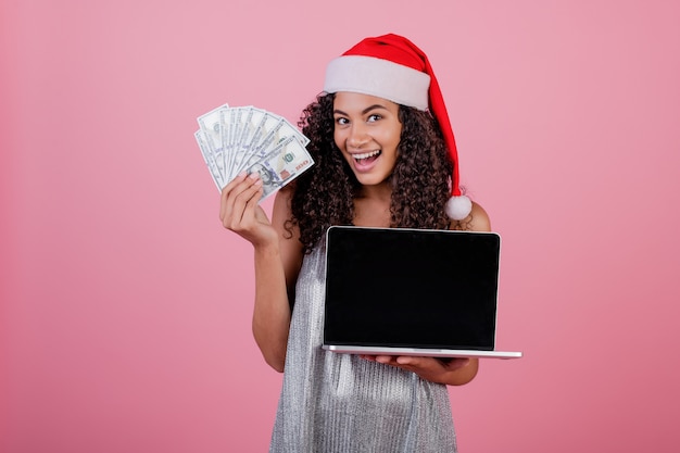 Donna di colore con lo schermo del copyspace sul computer portatile e denaro contante che porta il cappello di Santa isolato sopra il rosa