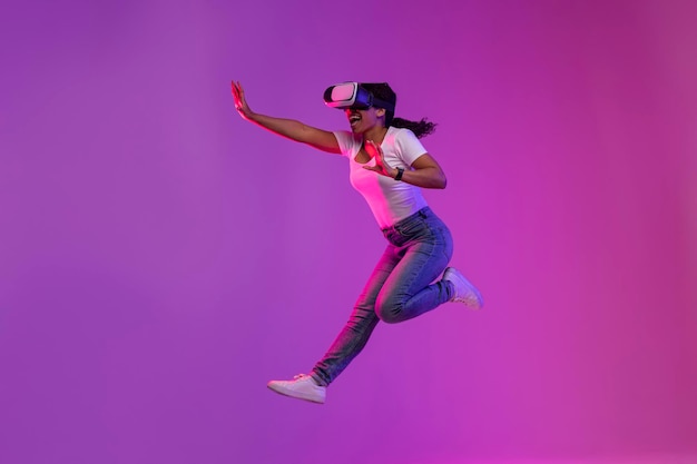 Donna di colore eccitata che indossa occhiali VR che saltano in aria alla luce al neon