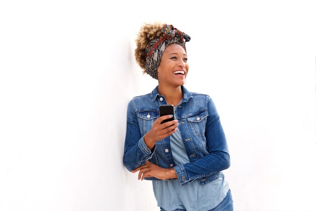 Donna di colore elegante in piedi sul muro bianco con il telefono cellulare e sorridente