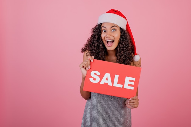 Donna di colore emozionante che tiene il segno rosso di vendita del copyspace e che porta il cappello di Santa isolato sopra il rosa