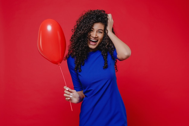 Donna di colore emozionante con l'aerostato a forma di cuore che porta vestito blu sulla parete rossa