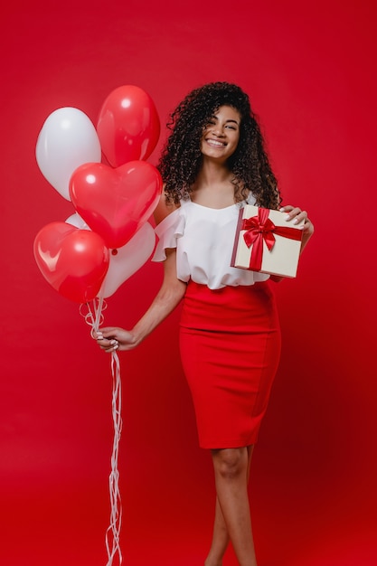 Donna di colore emozionante con palloncini colorati a forma di cuore e regalo di San Valentino sul muro rosso