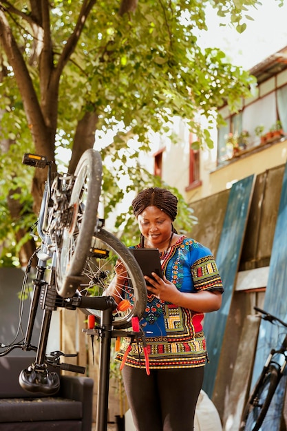 Donna di colore energica ed entusiasta che ripara e mantiene diligentemente vari componenti della bici utilizzando la tavoletta digitale nel cortile di casa. Giovane donna sportiva che naviga in internet per mantenere la bicicletta.