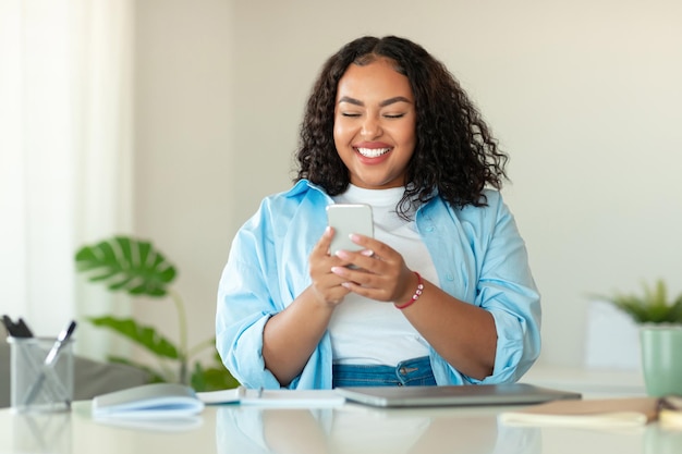 Donna di colore felice che utilizza il telefono cellulare che si siede nell'ufficio moderno
