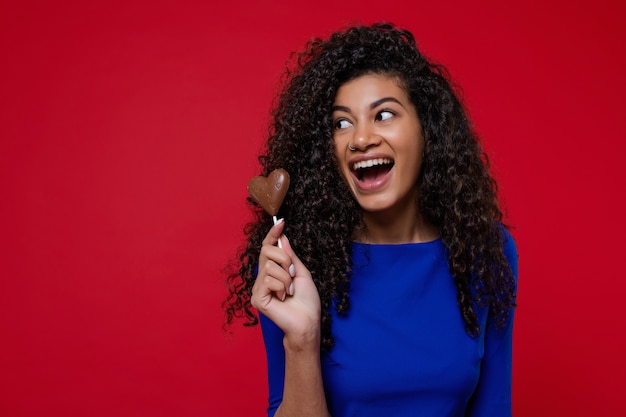 Donna di colore felice con la caramella di cioccolato a forma di cuore che sorride sulla parete rossa
