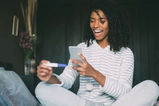 Donna di colore felice con un test di gravidanza e uno smartphone sul letto