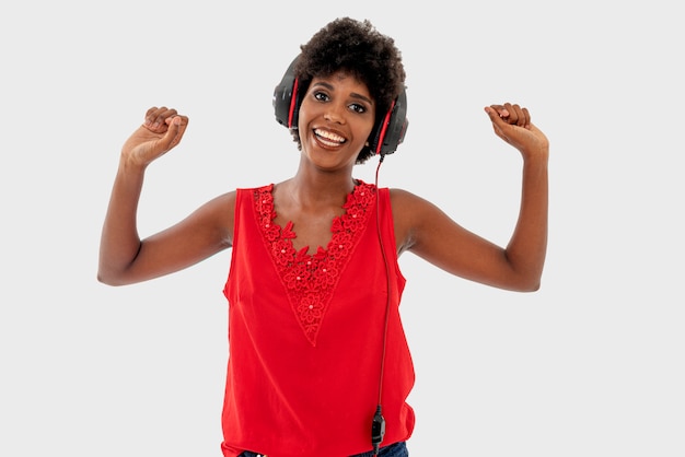 Donna di colore in camicia rossa isolata sopra fondo bianco, cantando e divertendosi mentre ascoltando la musica usando le cuffie