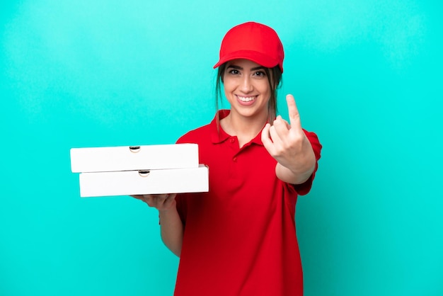 Donna di consegna della pizza con uniforme da lavoro che raccoglie scatole di pizza isolate su sfondo blu facendo un gesto in arrivo