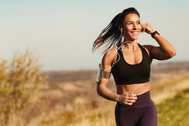 Donna di corporatura muscolare felice che ascolta musica con gli auricolari mentre corre in natura Spazio di copia